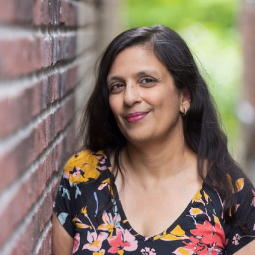 Sonal Champsee - author picture. A South Asian woman with long wavy hair, and a slight smile, wearing a black floral dress, standing next to a red brick wall